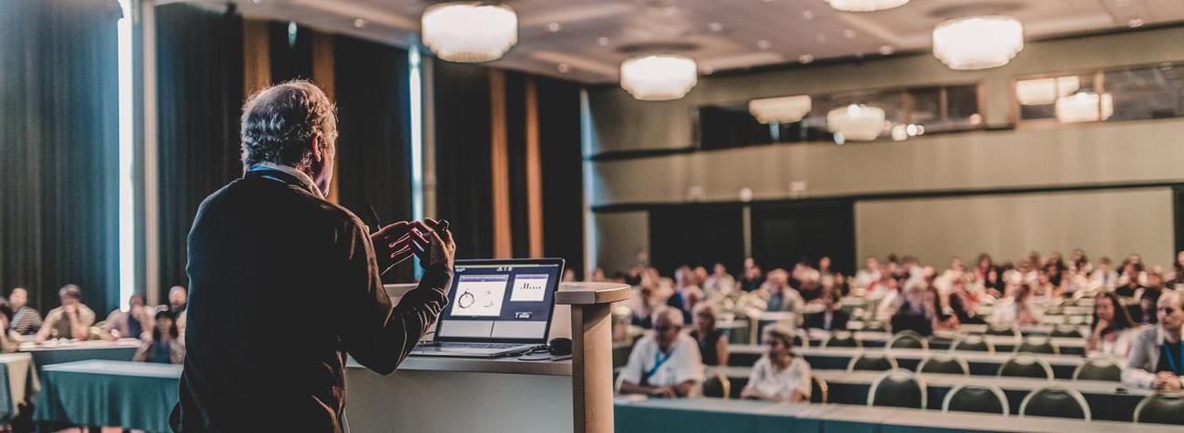 A professor stands in front of a podium while lecturing to a classroom full of people